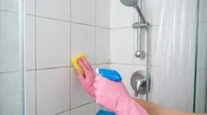Persona pulisce le piastrelle della doccia con guanti rosa, spugna e spray