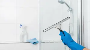Mano con guanto blu pulisce il vetro della doccia con una spatola tergivetro